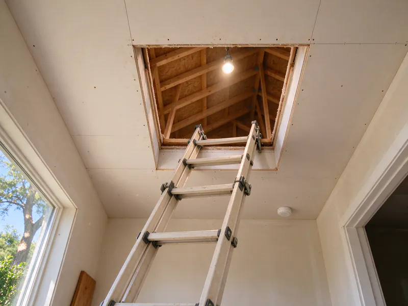 A ladder leaning up into an open attic access in the ceiling of an Austin house, a work light lit in the attic above