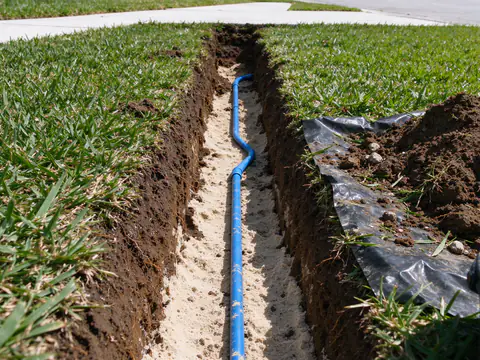 Open trench across a front lawn in Austin with a new blue PEX water service line bedded in sand, running from the curbside meter box toward the house